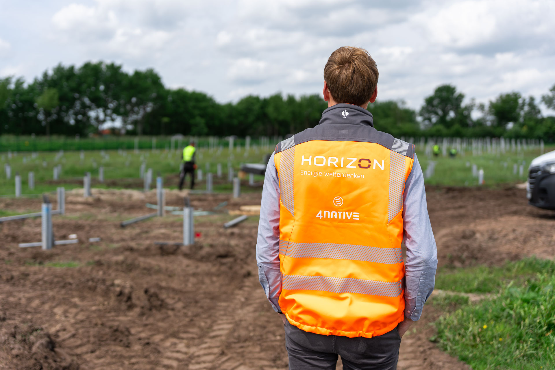 HORIZON-Geschäftsführer Thomas Kals trägt eine orangefarbene Warnweste mit 4native- und HORIZON-Logo. Er steht mit dem Rücken zur Kamera und schaut auf den Aufbau einer PV-Freiflächenanlage.