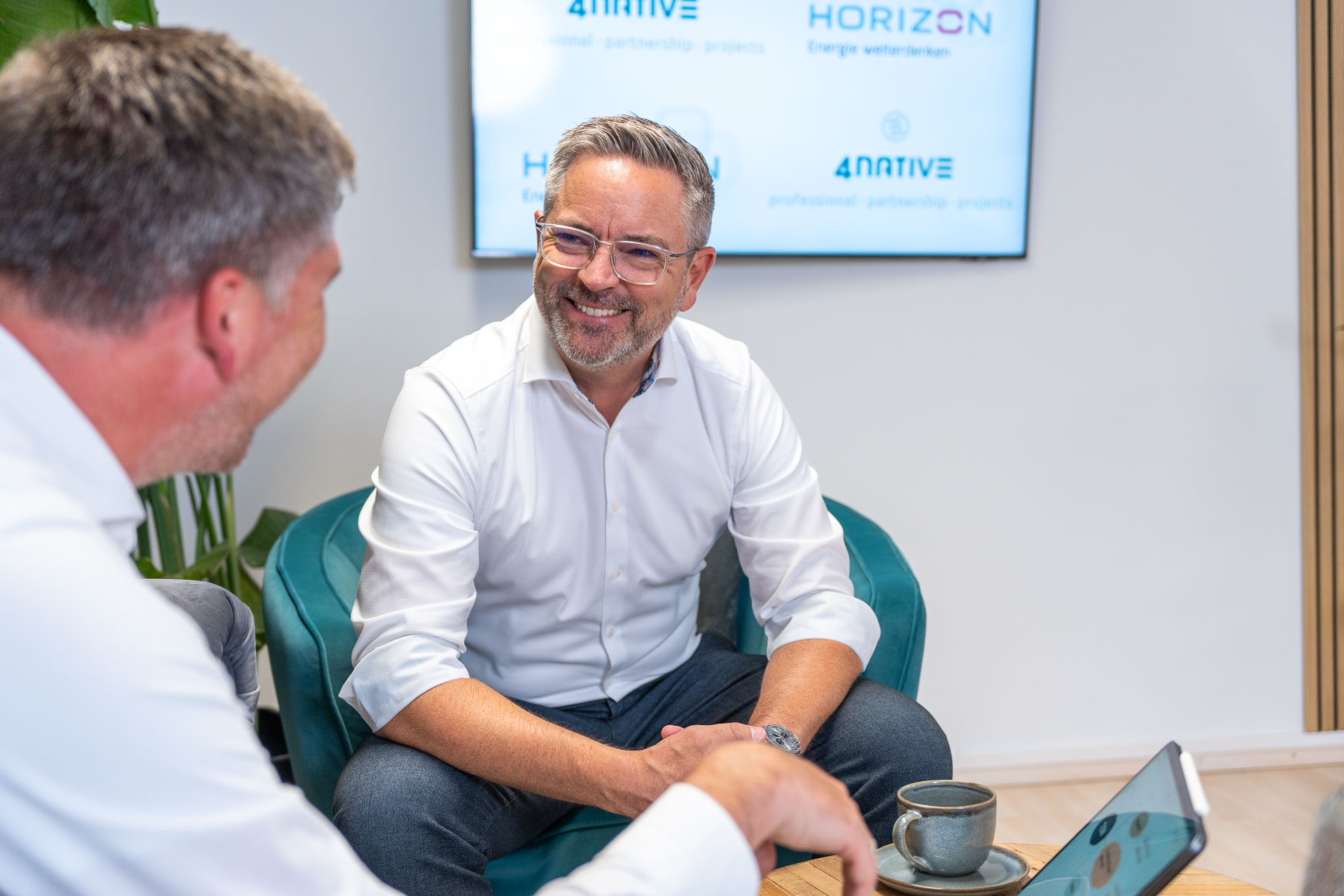 Zwei Mitarbeiter in weißen Hemden sitzen einander bei einem kleinen Tisch mit Laptop gegenüber und lächeln sich an.