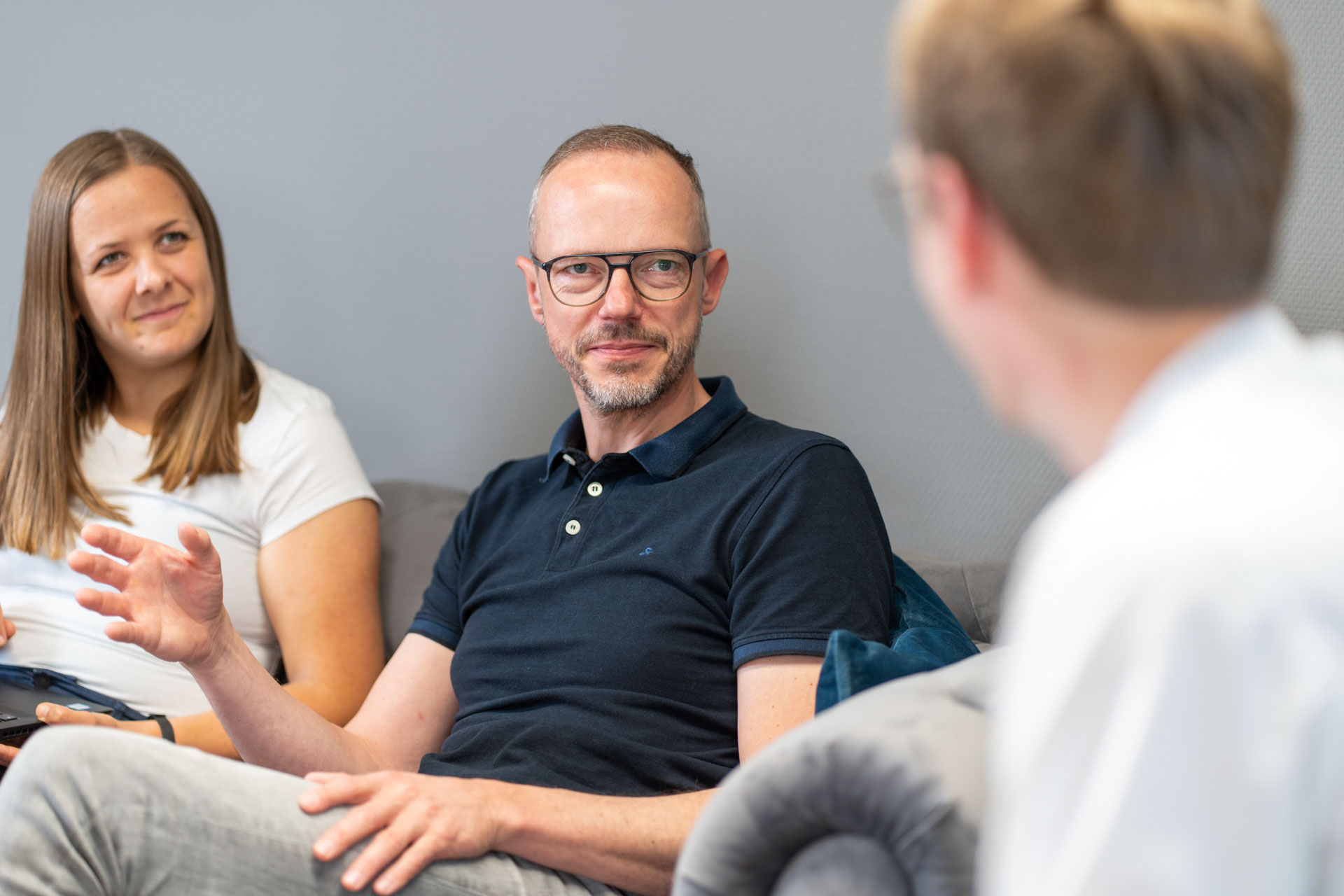 Eine Mitarbeiterin im weißen Shirt und ein Mitarbeiter im blauen Hemd sitzen dem HORIZON-Geschäftsführer gegenüber. Sie sind im Gespräch miteinander.