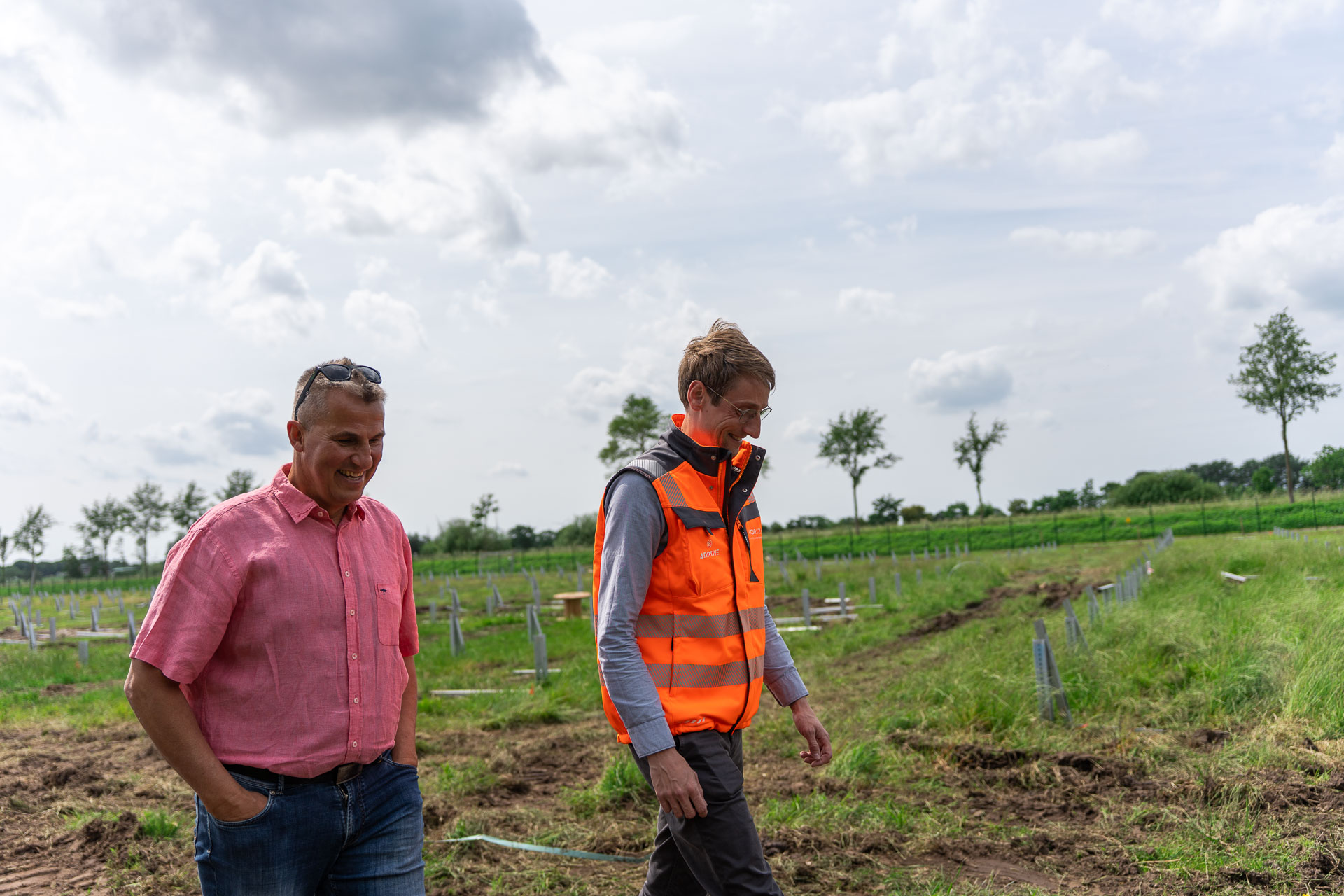 Ein Herr in pinkem Hemd und ein Herr in Warnweste gehen gutgelaunt nebeneinander über ein Feld.