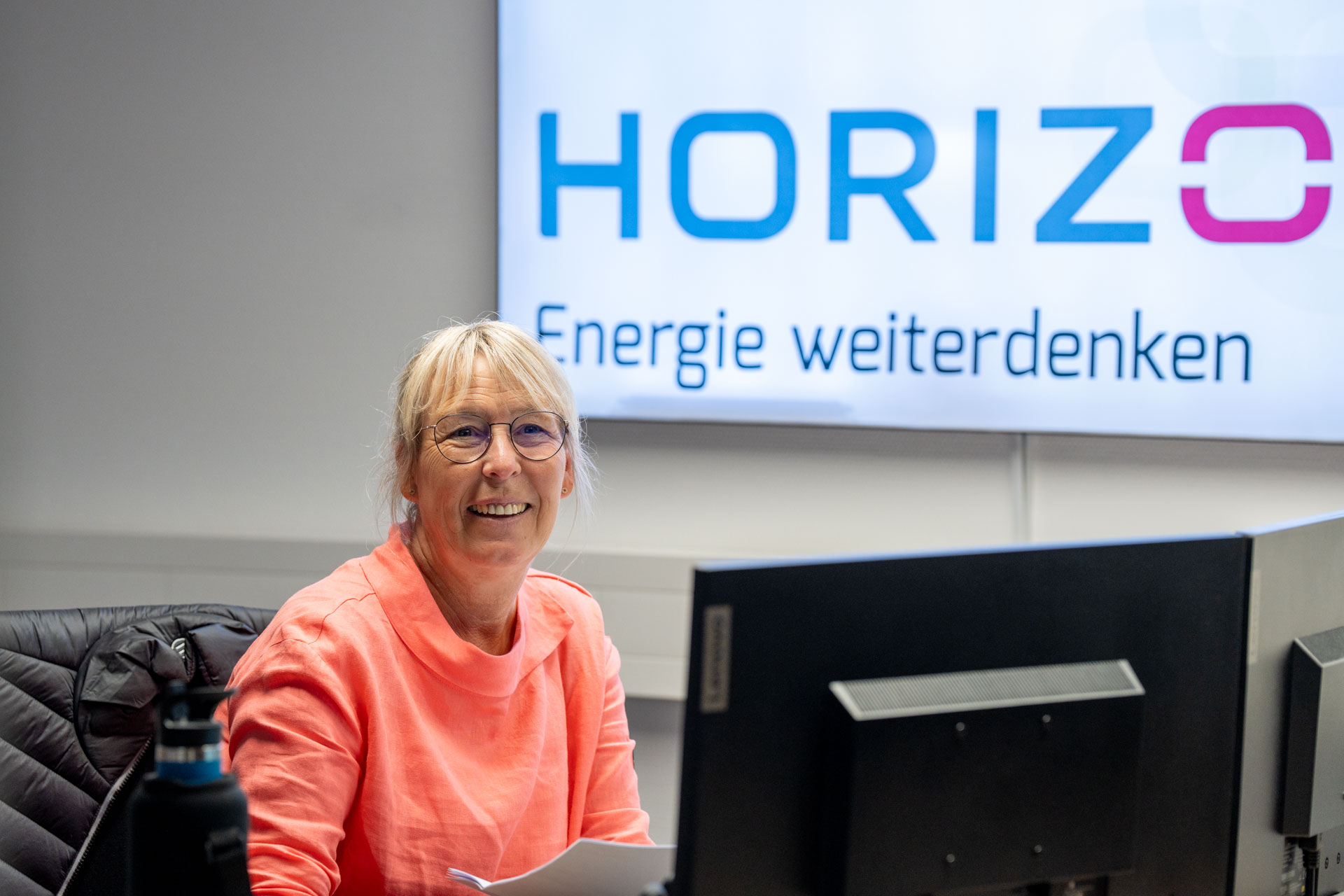 Eine freundlich lächelnde Frau sitzt hinter zwei Monitoren am Schreibtisch. Im Hintergrund ist groß das HORIZON-Logo auf einem Bildschirm.